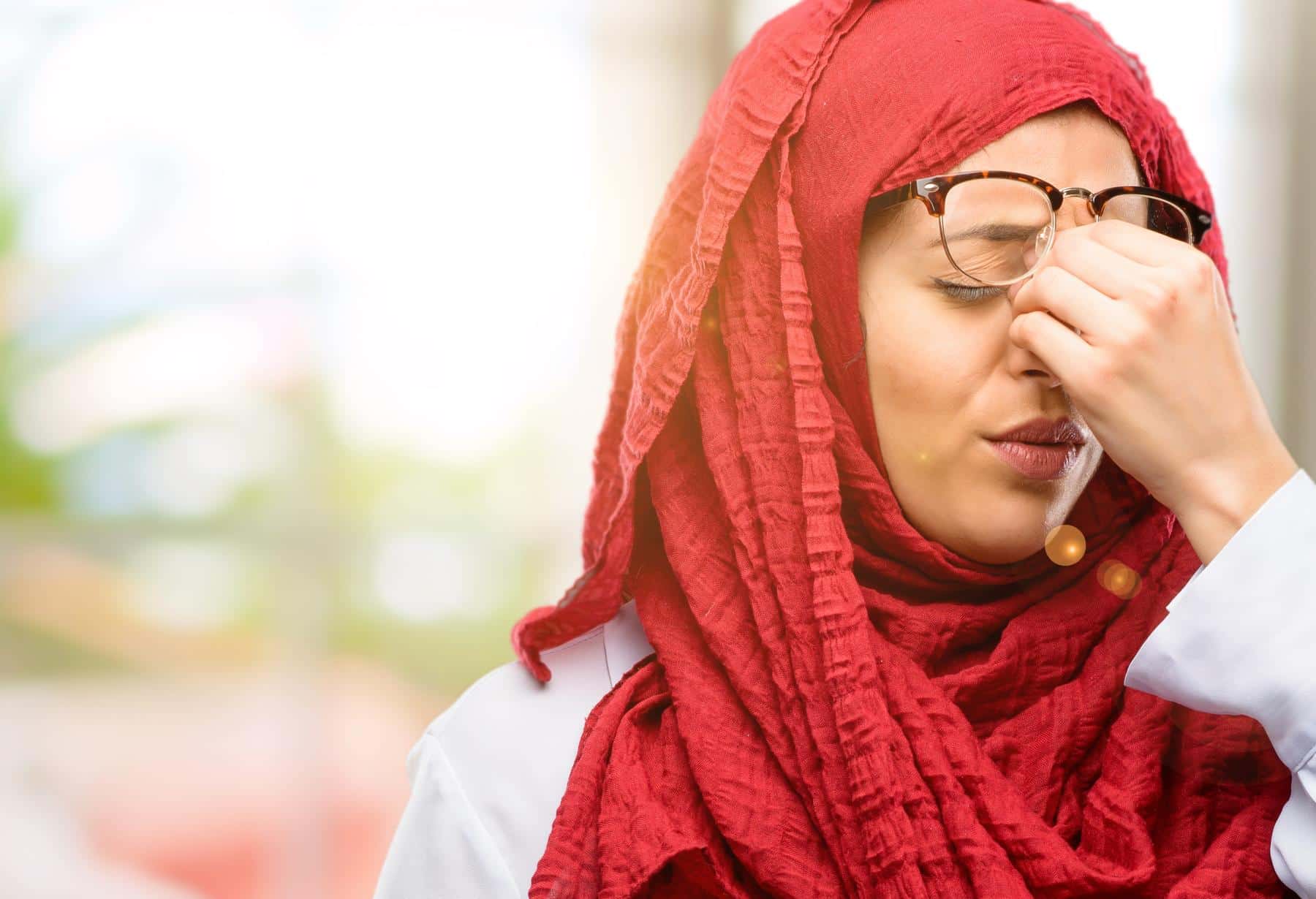 Woman in a red hijab pinching the bridge of her nose in pain, illustrating sar dard ki wajuhat