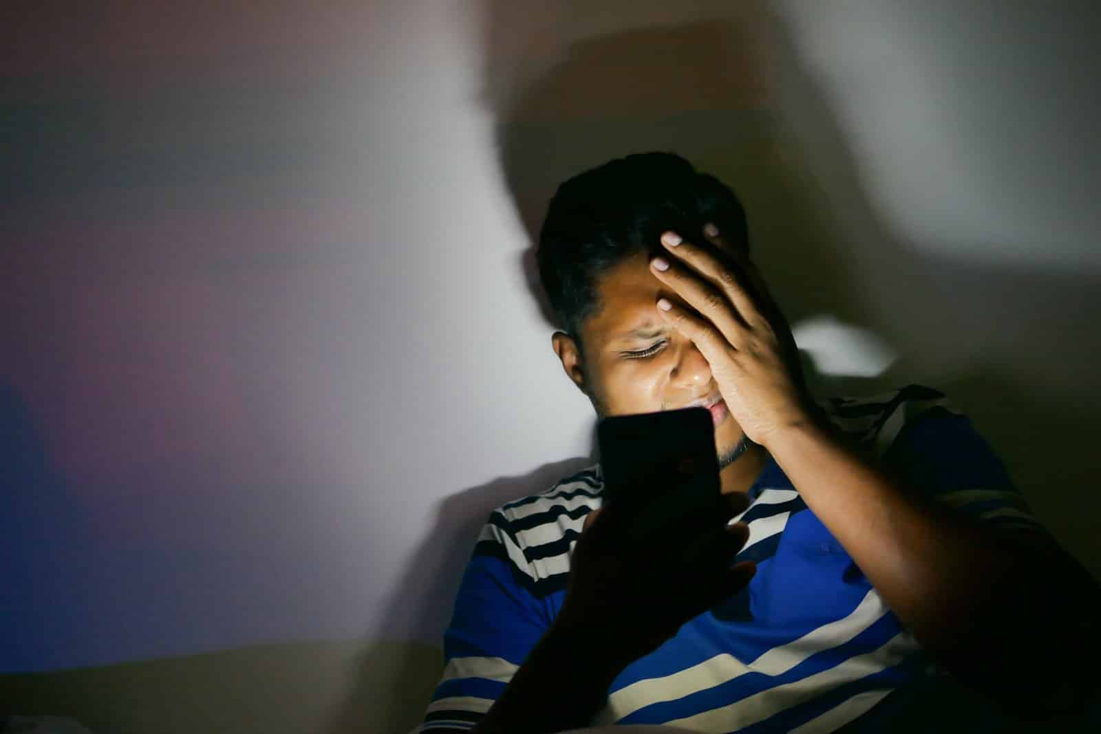 Man in a dark room lit only by his phone screen, holding his forehead in pain