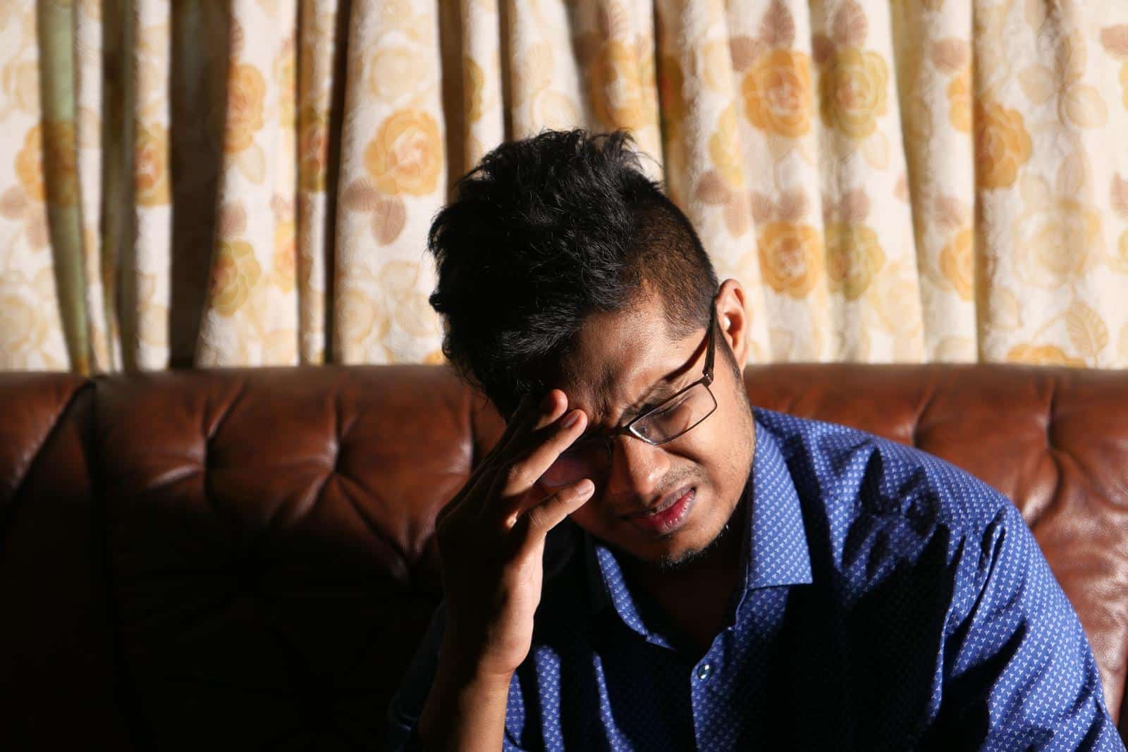 Man sitting on a couch at home holding his forehead, a common setting for skipped-meal headaches