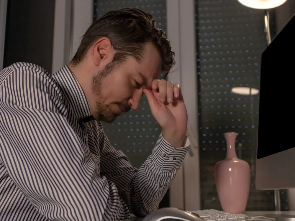 Man at his desk holding his forehead with eyes closed, showing headache and tiredness after a head injury