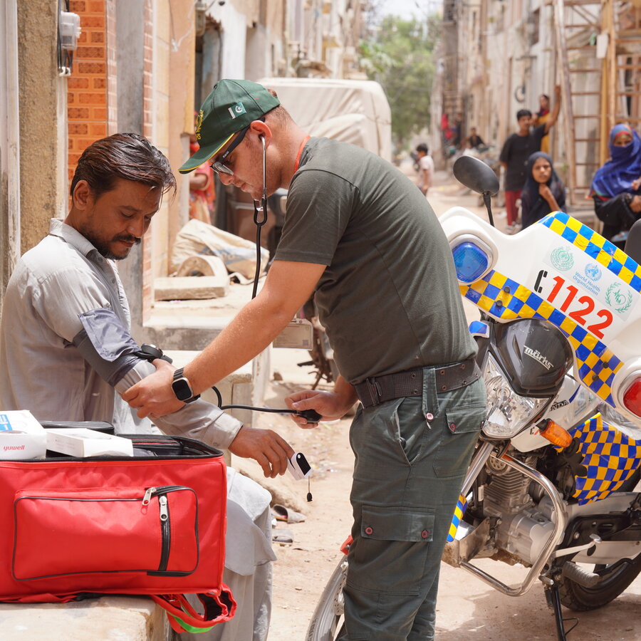 SIEHS 1122 paramedic checking a community member's blood pressure beside a branded motorbike ambulance in Karachi