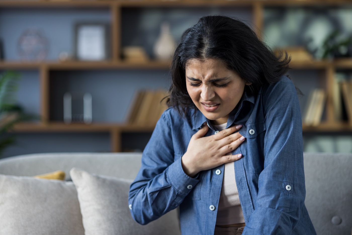 Young woman leaning over a couch with her hand on her chest, wincing in distress during a panic attack