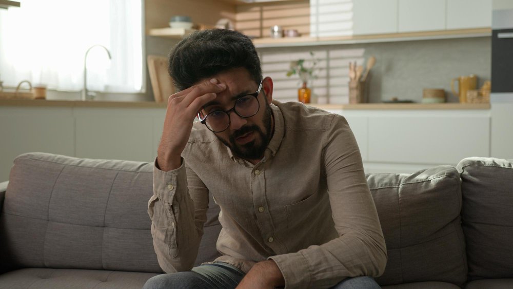 Man sitting on a couch at home with his hand on his forehead, looking anxious and overwhelmed