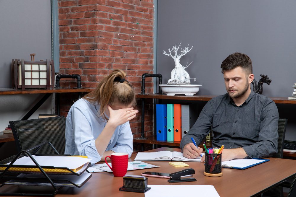 Two colleagues at a desk, one resting her head in her hand under workplace stress