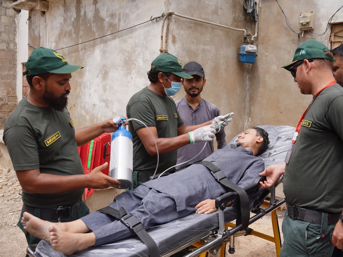 Three SIEHS paramedics preparing an oxygen mask and cylinder beside a patient on an ambulance stretcher in a street
