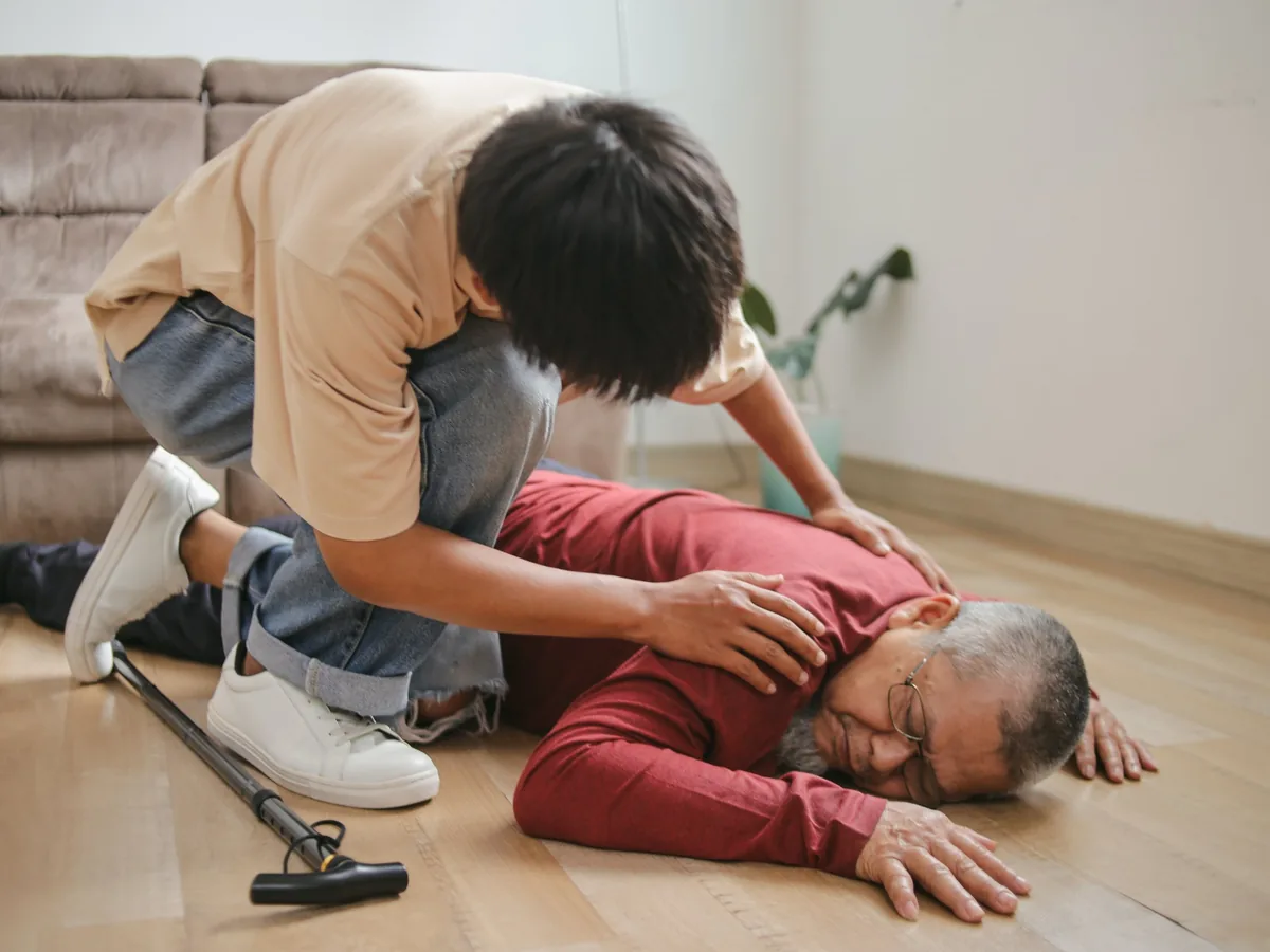 A young man kneeling on the living room floor with his hands on the shoulders of an older man who has fallen, a walking cane beside them
