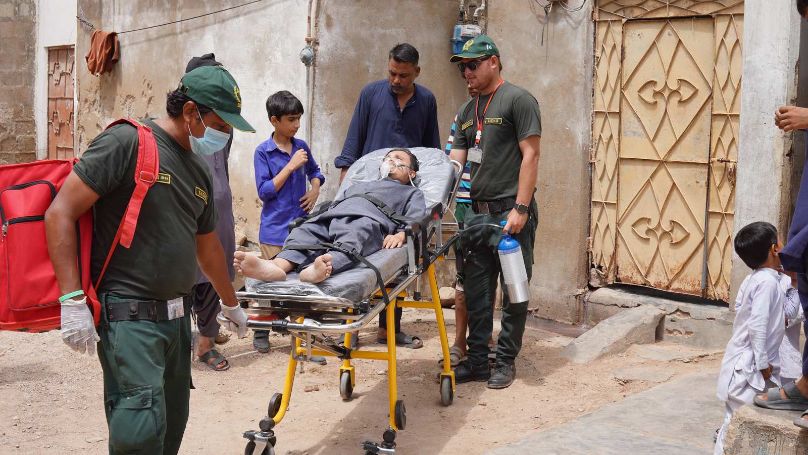 SIEHS 1122 paramedics wheeling a patient on oxygen on a stretcher in a Karachi neighbourhood
