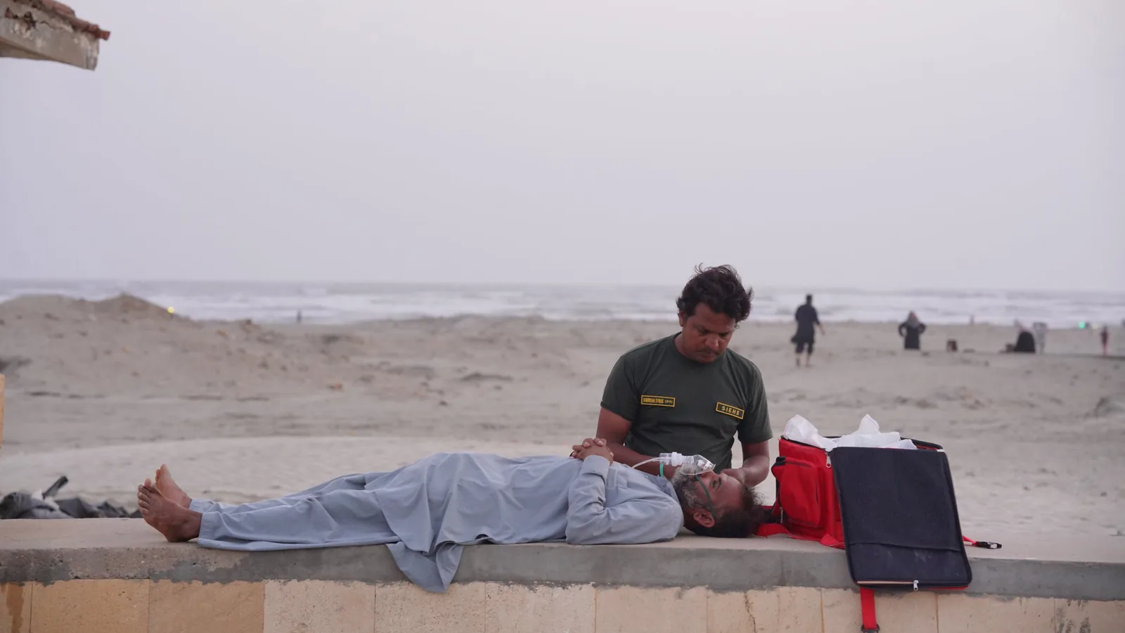 SIEHS paramedic kneeling beside a patient lying on a low wall on a beach, with an oxygen mask on and a red medical bag open beside them