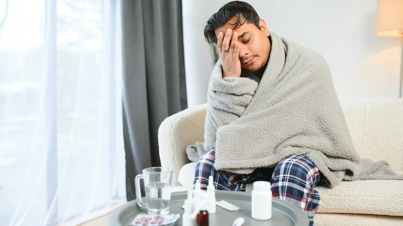 Typhoid symptoms, young man wrapped in blanket at home with medicines and thermometer on table